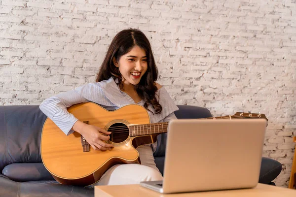 Mujer asiática joven practicando y aprendiendo a tocar la guitarra en el monitor del ordenador portátil. Guitarrista femenina viendo tutorial en línea —  Fotos de Stock