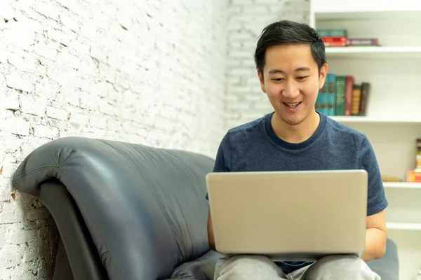 소파에서 평범 한 옷을 입고 소파에서 노트북 컴퓨터를 하고 있는 아시아 청년이 웃고 있습니다. 집에서 노트북으로 일하는 남성 프리랜서. — 스톡 사진