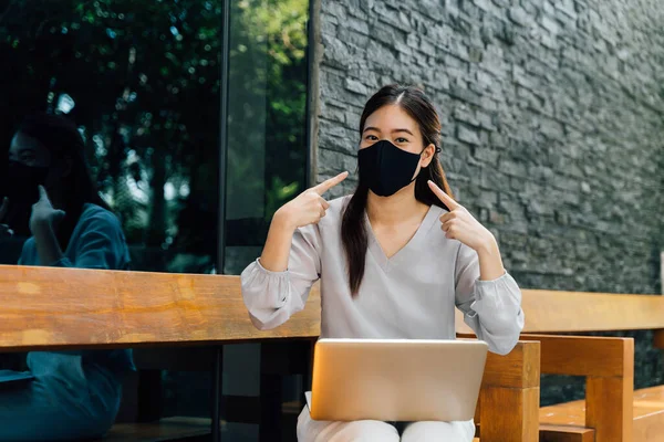 Mulher asiática usando uma máscara facial fora, enquanto sentado no laptop e pointin em sua máscara. Não se esqueça de usar uma máscara e máscara facial exigência para sair conceito — Fotografia de Stock