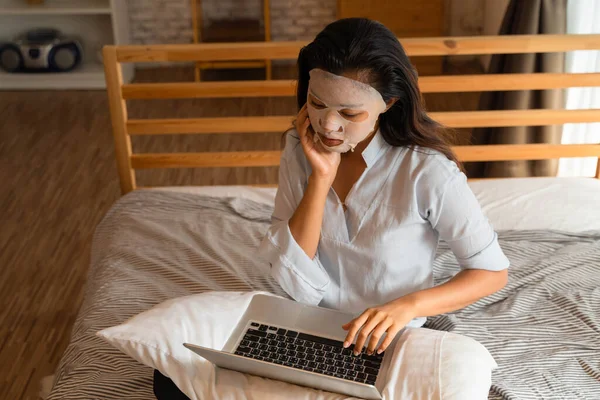 Retrato de una joven asiática que se aplica mascarilla facial en la cara mientras usa la computadora portátil en el dormitorio. Concepto de belleza y cuidado facial para mujeres —  Fotos de Stock