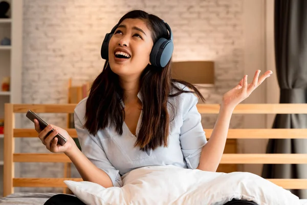 침대에 앉아서 집에서 휴대 전화를 들고 있는 동안 이어폰으로 노래하고 음악을 듣고 있는 행복 한 아시아 소녀 — 스톡 사진