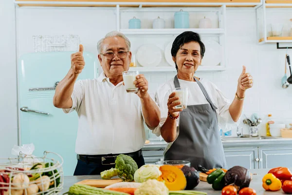 Szczęśliwa azjatycka para pije mleko w domu. 70-te Starszy żonaty mężczyzna i kobieta dając kciuki w górę i trzymając szklankę mleka w kuchni — Zdjęcie stockowe