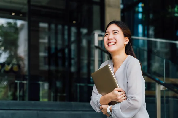 Seorang wanita bisnis remaja sukses dan bahagia memegang laptop dengan senyuman. Pekerja wanita profesional berdiri di kantor modern perusahaan dan jendela dalam tampilan abu-abu kasual. Dengan ruang fotokopi — Stok Foto