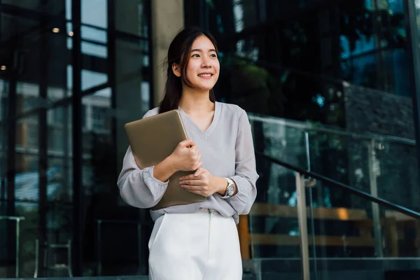 One successful and happy young adult business woman holding a laptop with smile. Professional female worker standing in corporate modern office and window in casual gray look. - With copy space — Stock Photo, Image