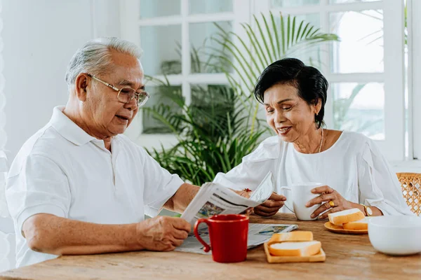 Starsza azjatycka para je razem śniadanie w pokoju obiadowym. Lata 70-te emerytowany mężczyzna i kobieta czytający gazety, cieszący się razem czasem. — Zdjęcie stockowe