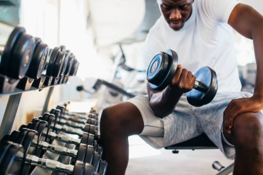 Genç Afrikalı Amerikalı bir adam spor salonunda oturmuş, halterini kaldırıyor.