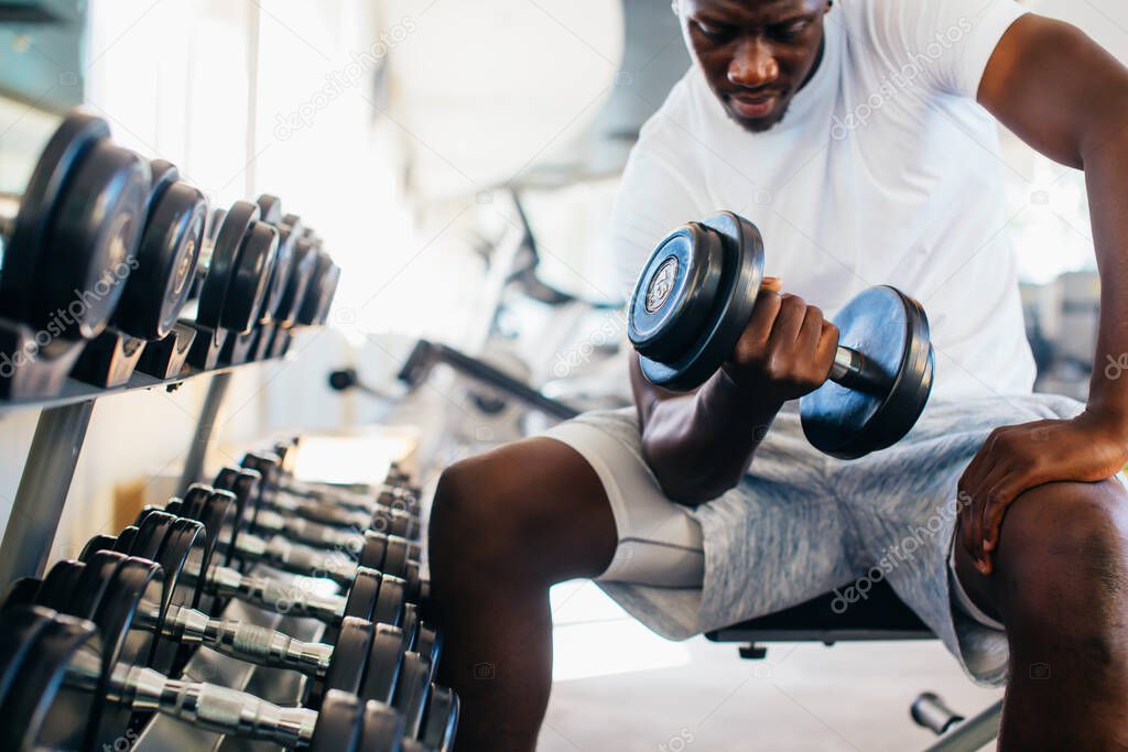 Young African American man sitting and lifting a dumbbell with the rack at gym