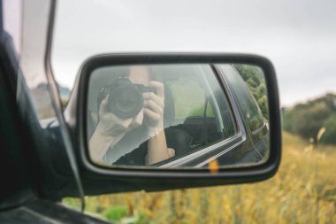Yolcu koltuğunda oturan sarışın kadın profesyonel fotoğrafçı dikiz aynasında sisli sarı dağ çayırlarının arka planına karşı selfie çekiyor.