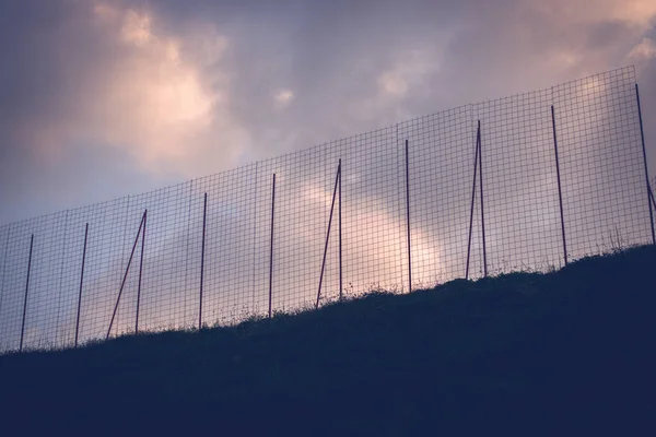 High metal wire net fence on hill against dark cloudy sunset sky