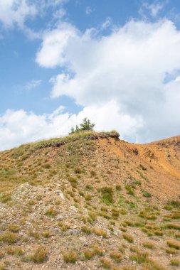 Beyaz bulutlara ve mavi gökyüzüne karşı çimlerle kaplı kayalık dağın tepesindeki kumlu plato.