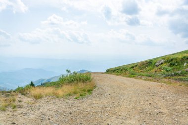 Kuru tozlu toprak yol bulutlu mavi gökyüzüne karşı yüksek uzak dağ sırasına nezaret ediyor.