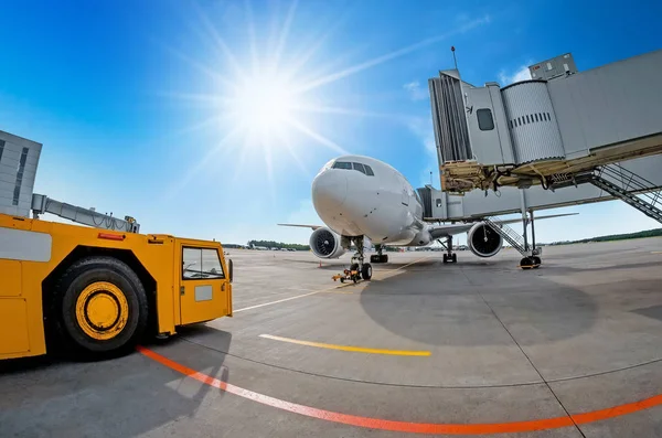 Aparcamiento en el aeropuerto, avión en la teletrampa. El tractor del aeródromo está listo para remolcar y salir de la aeronave. Sobre el fondo de un cielo azul y sol brillante, buen tiempo . — Foto de Stock