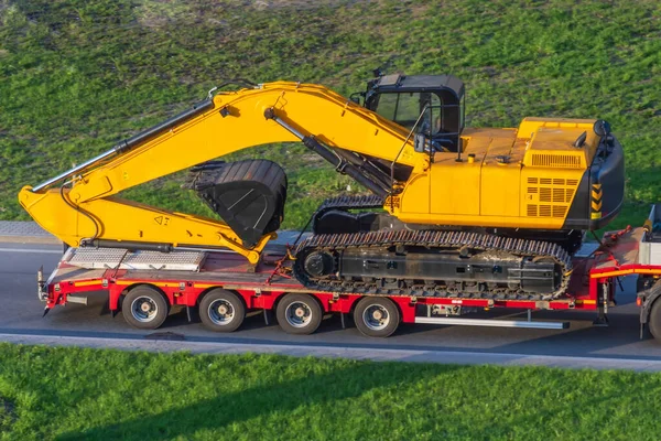 Heavy yellow excavator on transportation truck with long trailer red platform on the highway in the city