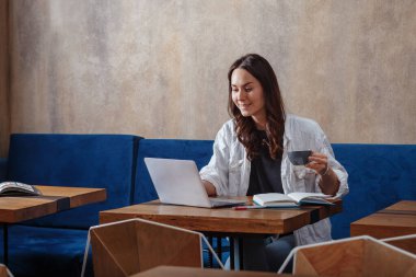 Bir kafede güzelce gülümseyen ve laptopuyla çalışan, kahve içen, not defterine not tutan genç bir kız.