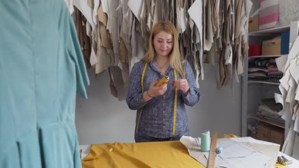 Модельер или швея, молодая женщина, выбирает цвет нитки к ткани и работает в студии, создавая стильную одежду. Уютная мастерская с оборудованием и инструментами, видимыми в — стоковое видео