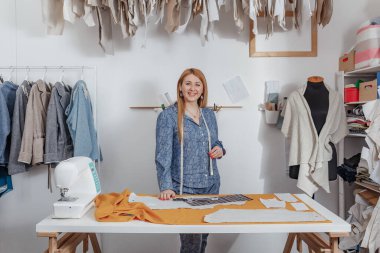 Mutlu genç bayan moda tasarımcısının portresi atölyede tek başına kameraya bakıyor. Küçük işletme sahibi ve başarılı gençlik konsepti.