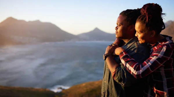 Casal inter-racial olhando para o pôr do sol vista para o oceano e montanha — Fotografia de Stock