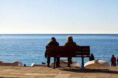 Aged couple, sitting on a bench in front of the sea, along the promenade of Camogli, take the winter morning sunlight. Camogli is a small fisherman village on the shores of the Ligurian Sea. (Northern Italy); during summertime became full of tourists