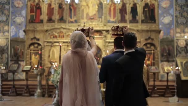 Ehrenrücken und Trauzeuge stehen hinter Braut und Bräutigam mit Kronen in den Händen über den verlobten Köpfen während der Hochzeitszeremonie vor dem Altar in der orthodoxen Kirche in schöner französischer Großaufnahme — Stockvideo