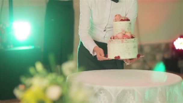 Garçom traz bolo de casamento e coloca-o na mesa sob luzes flash foto fechar sem rosto. Bonita decoração de bolo de casamento com flores de rosas naturais e bagas. Arranjo do banquete e restauração — Vídeo de Stock