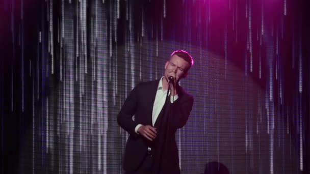 Man sings in microphone on stand at stage close up. Stylish handsome singer performs song with emotions on face at light LED screen background showing abstract animation of slowly falling rain lines — Stock Video