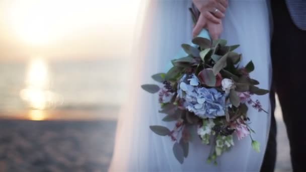 Bouquet di nozze insolito chiudere all'aperto. Le mani della donna tengono bouquet da sposa composto da fiori e foglie verdi al tramonto sullo sfondo vista mare. Decorazione floreale di design. Sposi in posa in spiaggia senza volto — Video Stock