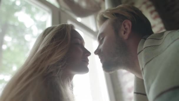 Kussen bij lichte venster backlit achtergrond sluiten van slow-motion. Jonge man komt om te kussen blonde vriendin op hem wacht thuis herfst ochtend weekend. Romantische paar relatie oprechte emoties — Stockvideo