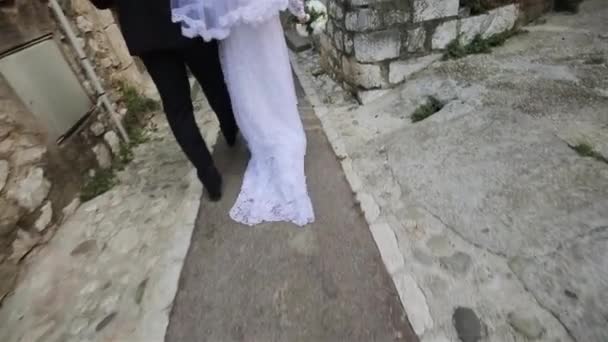 Newlywed couple bride and groom walk with only back seen down narrow street of ancient Italian or French town slow motion. Young wife wear white wedding dress with trail. Destination wedding in Europe — Stock Video