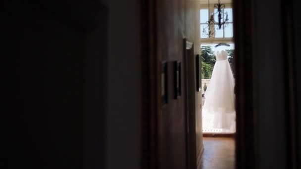 Abito bianco appendere alla porta della luce balcone in attesa di usura. Luce sera bianca o abito da sposa sul gancio visto dalla stanza corridoio buio. Bridal fashion design mattina di sposa estate armadio in onda — Video Stock