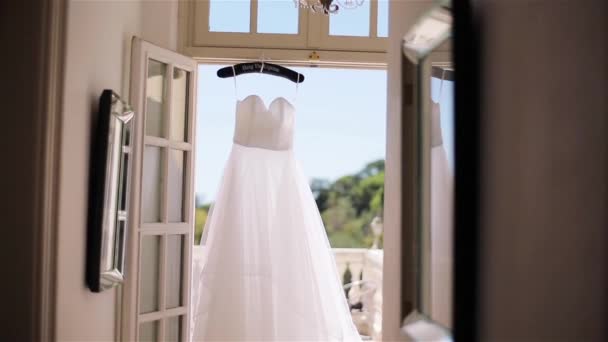 Das weiße Kleid hängt an einem speziellen Kleiderbügel in der Tür zur Terrasse aus nächster Nähe. Leichtes Sommerkleid aus Seiden- oder Chiffonstoffen wartet auf die Braut am sonnigen Morgenbalkon. Hochzeit Mode Schneiderei Designstudio — Stockvideo