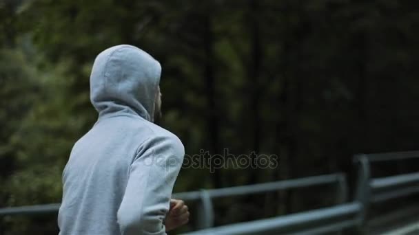 Homem correndo na montanha estrada serpentina até nenhum lado do rosto visão traseira close up câmera lenta. Corredor com capuz de manhã em cinza desgaste e tênis funciona exercitando treinamento ao ar livre. Esforço estilo de vida saudável — Vídeo de Stock