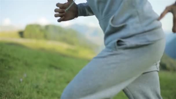 El hombre que se calienta en la naturaleza corre jogs cuesta arriba al aire libre en el campo cerca de la cámara lenta poco profunda. Persona encapuchada en ejercicios de ropa deportiva gris trabajando en hierba verde de la mañana. Estilo de vida saludable activo — Vídeo de stock