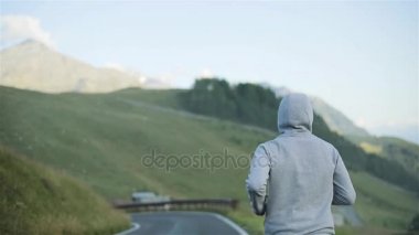 Doğada kırsal dağ yolda geri koşu adam ağır çekim kadar kapatın. Kapşonlu erkek figürü çalıştırmak sabah tepenin yamacında arka planda gri spor giyim ve spor ayakkabı yerel asfalt yolda takip