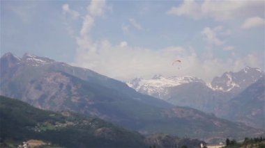 Dağ manzara uzak mesafe yamaç paraşütü. Yüzmek için turuncu katıyağ kullanarak bir Yamaçparaşütü uçmak ağaçlar güneş kar kepli doruklarına yaz video profili Valley çember yavaş düşük inerler. Hava spor turizmi