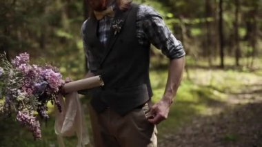 Otantik iyi giyimli damat kağıt rulo mektup parsel kır çiçekleri buketi doğa arka plan ağır çekim tutar. Çekici sarışın adam Festival kıyafeti sana ormanda orman sığ dof metinde yürür