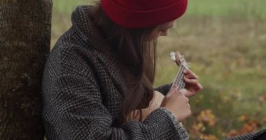 Hippi kızın ukulele ile birlikte doğa arka planında oturduğu yakın plan portresi. Kırmızı şapkalı genç kadın Hawaii gitarını yavaş çekimde çalmayı öğreniyor. Müzik eğitimi.