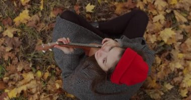 Sonbahar parkının arka planında ukulele çalan genç bayan müzisyen portresi. Akustik müzik aleti tutan mutlu bir kadın sarı kuru yapraklarla oturuyor.