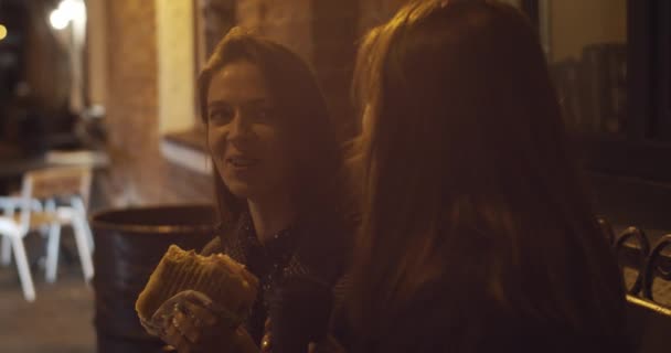 幸せな女性の友達と一緒にゆっくりと動きの外の通りの食べ物を食べる楽しい時間を過ごす 2人の若い女の子が市内のカフェでチーズバーガーコーヒー1泊黄色の光を保持座って笑う 不健康な食事 — ストック動画