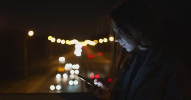 Parlak trafik ışığı geçmişinde akıllı telefon arayan genç bir kadının yan portresi. Geceleyin elinde daktilo tutan çekici bir kız ve arkasında da çözülmüş bir bokeh var. Akıllı teknolojiler sosyal medya