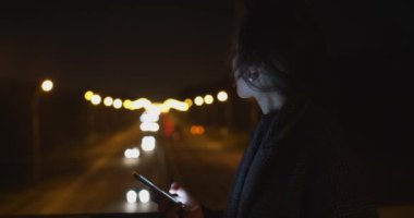 Geceleri dışarıda cep telefonu kullanan güzel bir kadın trafik ışıklarının arkasını değiştiriyor. Yavaş çekimde akıllı telefondan mesaj gönderen bir kızın doğal portresi. Gece yoğun yaşam tarzı aktiviteleri