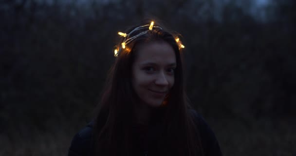 ガーランドライトが燃える美しい女性は 夕暮れの自然を背景に屋外でポーズをとっています 森の中で幸せな笑顔のお祝いの装飾冠花輪のモデルの自然な肖像画 — ストック動画