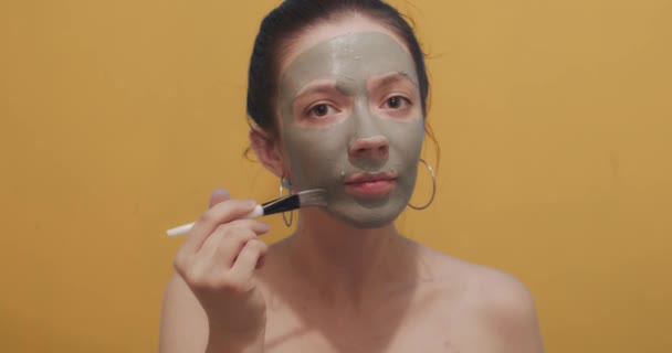 化粧ブラシを使用して顔マスクを適用パンと自然なブルネットは黄色の背景をモックアップします 屋内スローモーションコピースペースをスキンケアを取って若い女性のフロント肖像画 女性美容室 — ストック動画