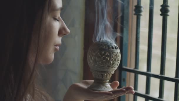 Side portrait of young woman smelling white fume of indian censer inside temple. Pensive girl meditating with closed eyes by burning incense smoke indoors slow motion close up. Mind body spirit — Stock Video