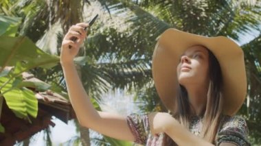 Neşeli bayan yeşil palmiye ağacının arka planında cep telefonuyla selfie çekiyor. Kendini tropikal doğada yavaş çekimde vuran mutlu kadın turistin portresi. Akıllı teknoloji uygulaması
