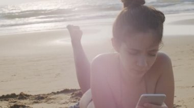 Sıska kız, bikinili, kumlu sahilde güneşleniyor. Elinde cep telefonu, açık hava portresi var. Deniz kenarında uzanan seksi kadın akıllı telefon yazıcı aygıtı yavaş çekimde kopyalama alanı metnini kapat
