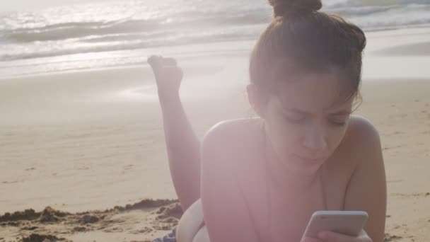 Schlankes Mädchen im Bikini sonnt sich am Sandstrand und hält ihr Handy hoch. Sexy Frau liegt am Meer Ozean eingeben Smartphone-Handheld-Gerät Zeitlupe Nahaufnahme kopieren Weltraumtext — Stockvideo