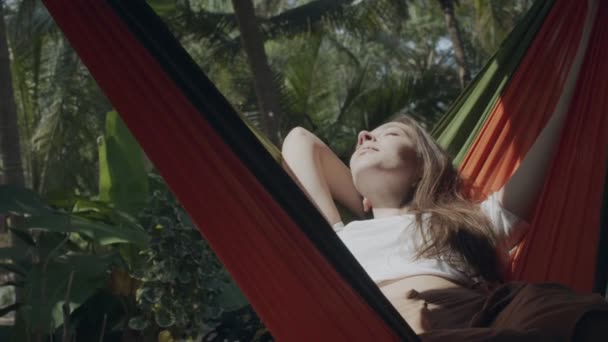 年轻女子日光浴躺在公园绿林的吊床上。迷人的女孩在阳光明媚的夏日里闭着眼睛放松自己。旅行健康的生活方式幸福的医疗保健 — 图库视频影像