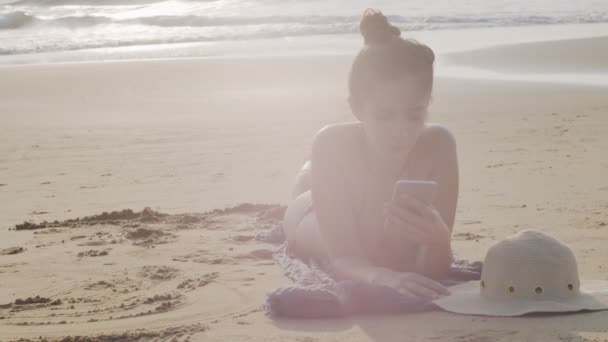 Femme attrayante paresser au soleil lecture fil d'actualité sur fond de rivage de l'océan smartphone. Bain de soleil touristique féminin positif sur la plage tenant le téléphone portable dans l'espace de texte de copie de lumière naturelle. Voyage été — Video