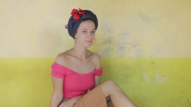 Sarı duvarda duran boho elbiseli türbanlı şık bir kızın portresi. Etnik saç modelli bir kadının açık havada ağır çekimde oturması ön planda. Tasarımcı şık kıyafetler yaz tatilinde seyahat eder.