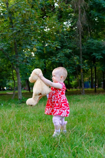 Uma menina brinca com um ursinho de pelúcia em um parque em uma grama — Fotografia de Stock